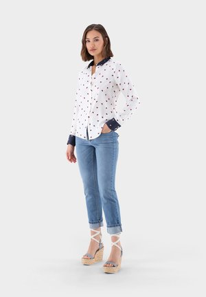 White shirt with red heart pattern, navy collar and cuffs. Paired with light blue jeans and tan wedge sandals.