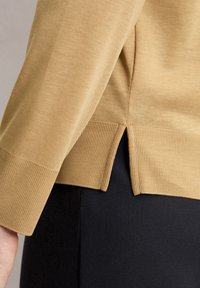 Beiger Pullover mit gerippten Manschetten und einem seitlichen Schlitz am Saum, der eine weiche Textur und eine taillierte Passform zeigt. Über einem schwarzen Kleidungsstück getragen.