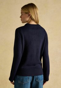 Marineblaues Strickpullover mit geripptem Kragen und Saum. Verfügt über lange Ärmel, eine lockere Passform und dezente Textur. Getragen mit hellblauen Jeans.