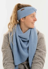 Donna con capelli biondi raccolti in una coda di cavallo che indossa una fascia per capelli blu a maglia e una sciarpa coordinata, sorridente e con lo sguardo rivolto a destra su uno sfondo neutro.