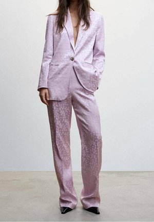 Femme portant un tailleur pantalon en satin violet clair avec des pois subtils, une main dans la poche, assorti à des chaussures noires à bouts pointus.