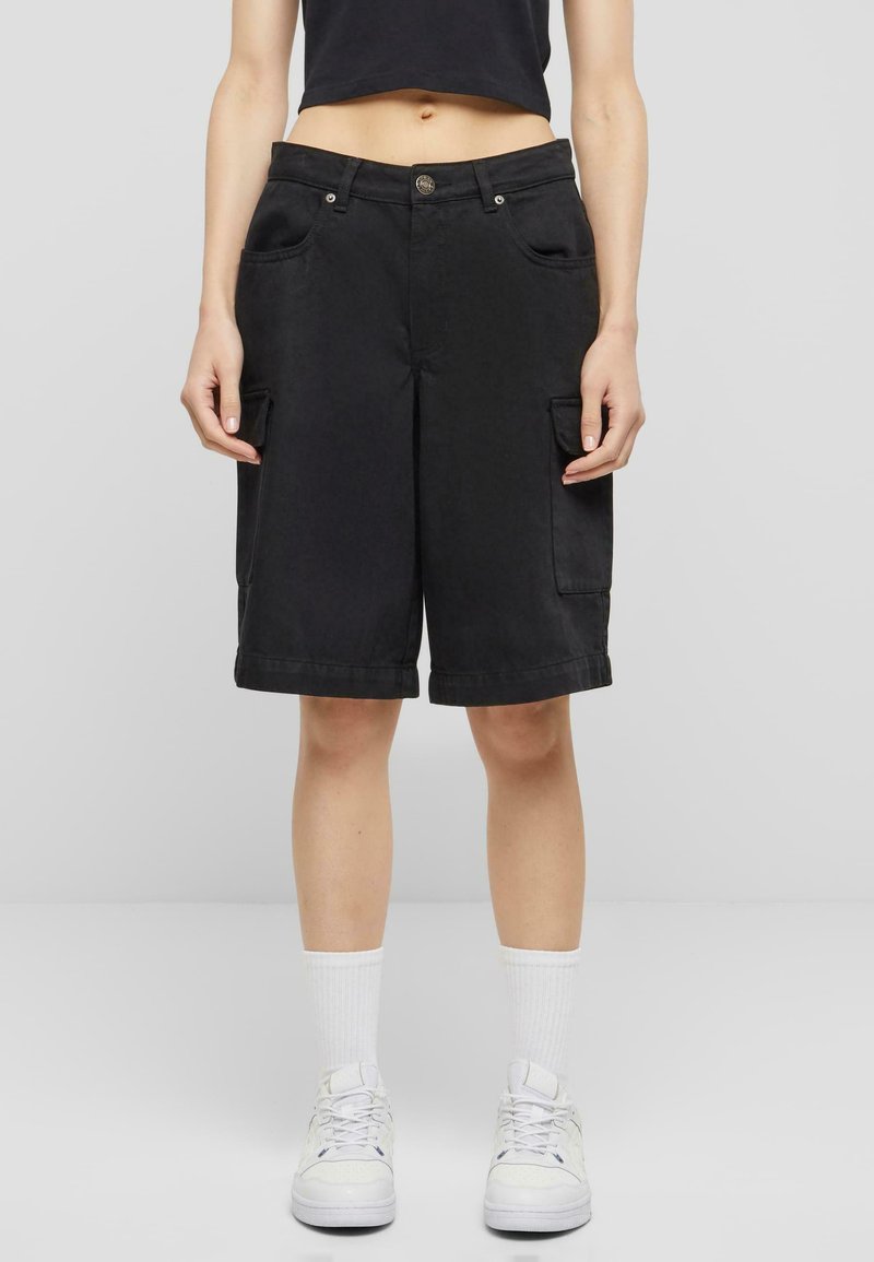 Kvinne iført svarte kne-lange cargo shorts med frontlommer, en svart kort topp, hvite sokker til ankelen, og hvite sneakers, som står på grått gulv.