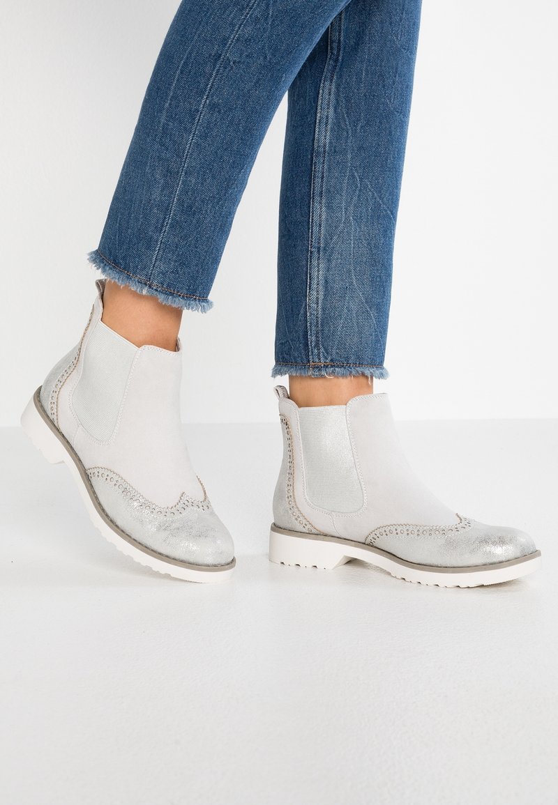 Bottes basses en cuir blanc avec embouts argentés et détails en aile portées avec un jean bleu à ourlet effiloché sur un sol blanc.
