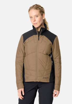 Blonde Frau trägt eine braun-schwarze Steppjacke mit Reißverschluss und schwarze Hose und steht vor einem einfarbigen hellen Hintergrund.