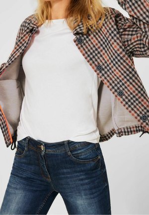 Kobieta ubrana w białą koszulę i niebieskie dżinsy, trzymająca otwartą brązowo-czarną kratowaną kurtkę z czarnymi guzikami.