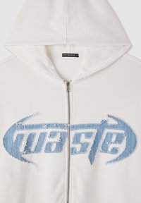 Felpa bianca con zip e un logo di denim blu sfilacciato sul petto, dotata di cappuccio. L'etichetta indica la taglia XL.