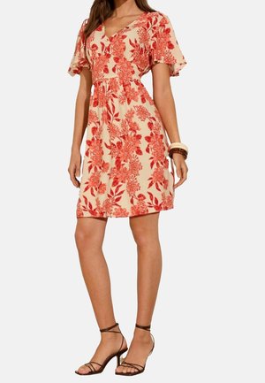 Femme portant une robe crème à manches courtes avec un imprimé floral rouge, associée à des sandales à talons hauts marron à brides et des bracelets à un poignet.