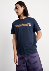 Jūras zilas kokvilnas T-krekls ar zeltu "Timberland" tekstu un logo. Vienkārša apaļa apkakle. Apvienots ar purpursarkaniem saistāmais šortiem.