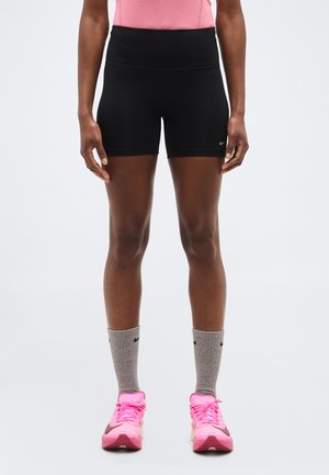 Person wearing black fitted shorts, gray crew socks, and bright pink athletic shoes, standing against a plain light background.