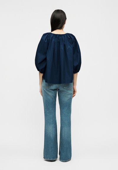 Γυναίκα με μακριά σκούρα μαλλιά, που φοράει μπλε μπλούζα με φουσκωτά μανίκια και ανοιχτόχρωμο καμπάνα τζιν, στέκεται με την πλάτη προς τα εμπρός σε απλό φόντο.