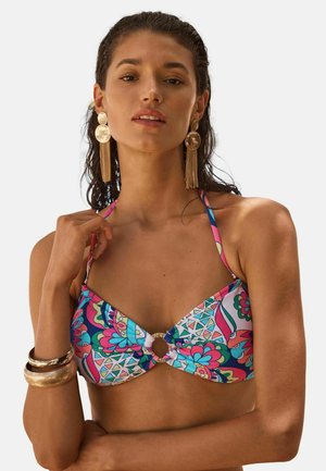 Vrouw draagt een kleurrijk bikini top met patroon, gouden creool- en kettingoorbellen en gouden armbanden, poserend tegen een effen achtergrond.