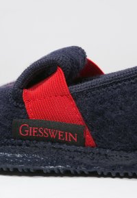 Navyblaue Wollslipper mit roten Stoffakzenten und einer strukturierten Sohle. Mit einem schwarzen Etikett, auf dem "Giesswein" in roten Buchstaben steht.