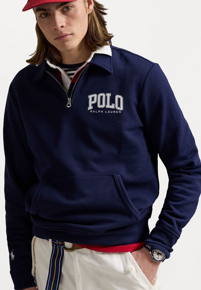 Marineblå pull-over sweatshirt med halvlynlås krave, med "POLO RALPH LAUREN" logo, frontlomme og ribbede ærmer. Hvid langærmet skjorte under.