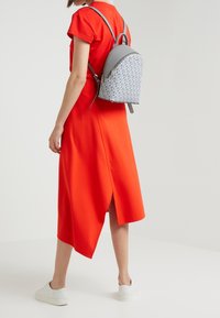 Grå mönstrad ryggsäck med rundad form, slät textur och justerbara remmar, bärs med en ljusröd klänning och vita sneakers.