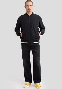 Juodas lengvas bomberis su užtrauktuku priekyje, raukšlėtomis rankovėmis ir apačia; dėvimas virš tamsių džinsų ir geltonų sportbačių su baltais akcentais.