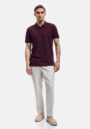 Junger Mann trägt ein bordeauxfarbenes Polohemd mit kurzen Ärmeln, hellbeige Hose und beige gewebte Slipper, steht vor einem einfarbigen Hintergrund.