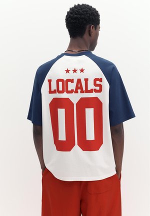 Mand iført en hvid og navy skjorte med "LOCALS" og "00" i stort rødt print på ryggen, kombineret med røde bukser, står med hænderne i lommerne.