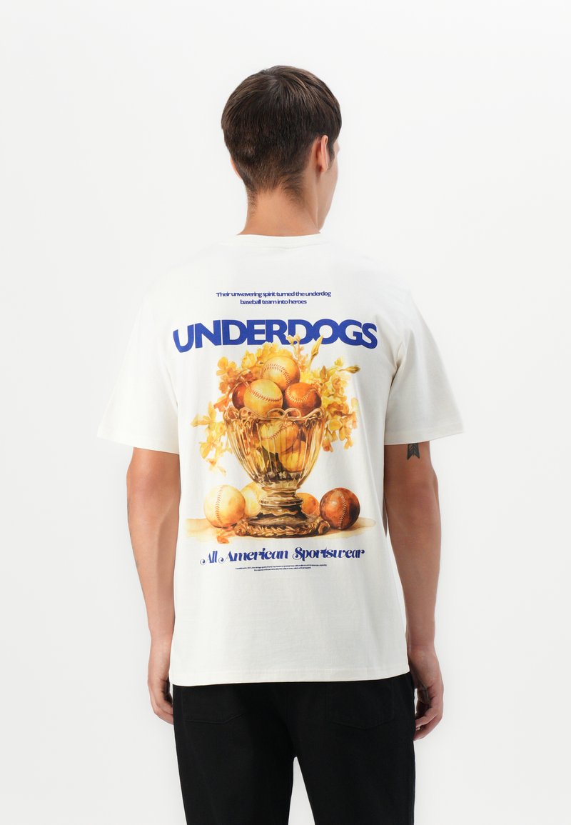 Mann som har på seg hvit T-skjorte med teksten "UNDERDOGS" og en illustrert trofé fylt med baseballer og blomster på baksiden, står mot hvit bakgrunn.