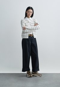 Cardigan alb cu model și dungi negre peste o cămașă cu guler, asortat cu pantaloni largi de culoare închisă și adidași maro.