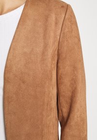 Gros plan d'une personne portant une veste en daim marron clair sur une chemise blanche côtelée, montrant la zone de l'épaule et du haut du bras.