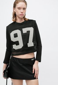 Schwarzes, kurzes Langarmoberteil aus Mesh-Stoff mit weißem "97"-Grafik, kombiniert mit einem schwarzen Minirock mit einem Marken-Patch.