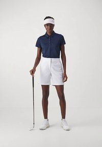 Marinblå pikétröja med knappar, vita skräddarsydda shorts, vit keps och vita sneakers; håller i en golfklubba. Enkel och funktionell design.