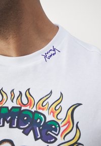 Biela bavlnená tričko s farebnými grafikami plameňov a textom "MORE". Výrazné fialové vyšívanie na golieri pridáva na detailoch.