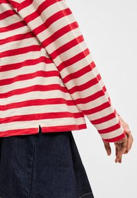 Sarkana un krēmīgi svītraina garās piedurknes blūze ar noapaļotu apakšdaļu un sānu slīpām. Valkāta virs tumši zila džinsu svārkiem, izceļot mīksto auduma tekstūru.