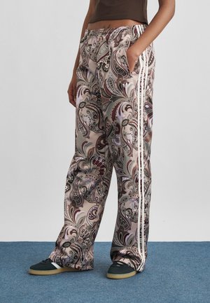 Persoon draagt wijde broek met paisley-patroon en witte zijstrepen, zwarte sneakers en een bruin cropped top, staand op blauw tapijt.