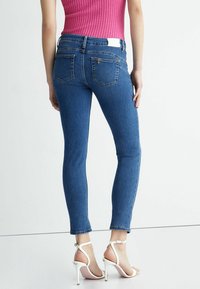 Mujer con jeans ajustados recortados azules, top sin mangas de punto rosa y sandalias blancas de tacón alto, de pie contra un fondo liso.