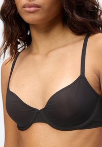 Γυναίκα που φοράει μαύρο σουτιέν με μπανέλα και λεπτές ρυθμιζόμενες τιράντες με την επωνυμία Calvin Klein σε απλό φόντο.