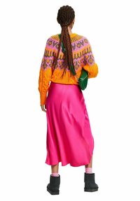 Pull en tricot orange vif avec motifs roses et violets, associé à une jupe midi en satin rose et des bottines noires. Sac à épaule vert.