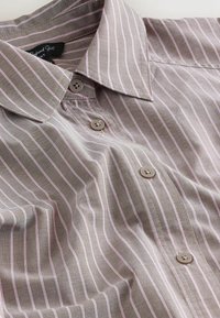 Košeľa na zapínanie na gombíky svetlohnedej farby s jemnými ružovými pruhmi. Má špicatý golier a tri hnedé gombíky. Hladká textúra látky.