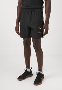 Los pantalones cortos deportivos negros cuentan con una cintura elástica y un logo de Puma amarillo en el lado izquierdo. Se llevan con zapatillas negras y naranjas.