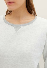 Sudadera gris clara con cuello de canalé, tejido texturizado y detalle de costura en V en el collar. Presenta un corte relajado y mangas largas.