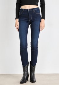 Tmavo modré skinny džínsy s minimalisticým dizajnom, vysokým pásom, akcentovaným prešívaním a predným zapínaním na gombík, skombinované s čiernymi podpätkovými čižmami.