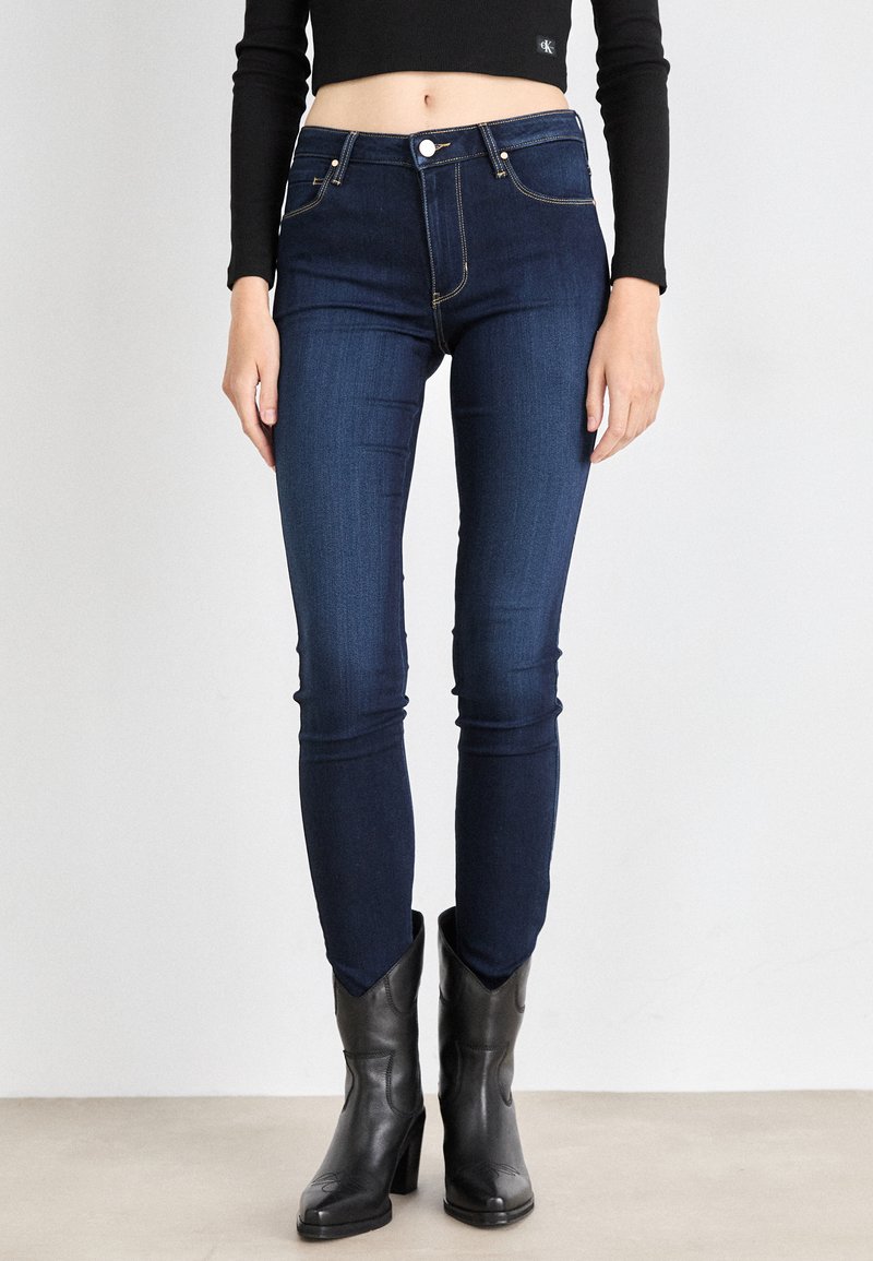 Tmavo modré skinny džínsy s minimalisticým dizajnom, vysokým pásom, akcentovaným prešívaním a predným zapínaním na gombík, skombinované s čiernymi podpätkovými čižmami.