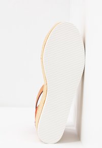 Semelle de sandale compensée avec dessous blanc texturé, semelle intermédiaire en liège et brides en cuir marron, positionnée verticalement contre un coin blanc.