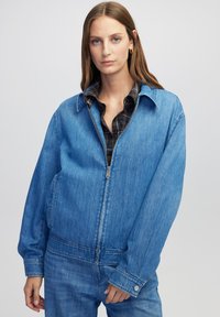 Jonge vrouw in een oversized blauwe spijkerjack en bijpassende jeans, met een gedeeltelijk dichtgeritste voorkant en een geruit shirt eronder.