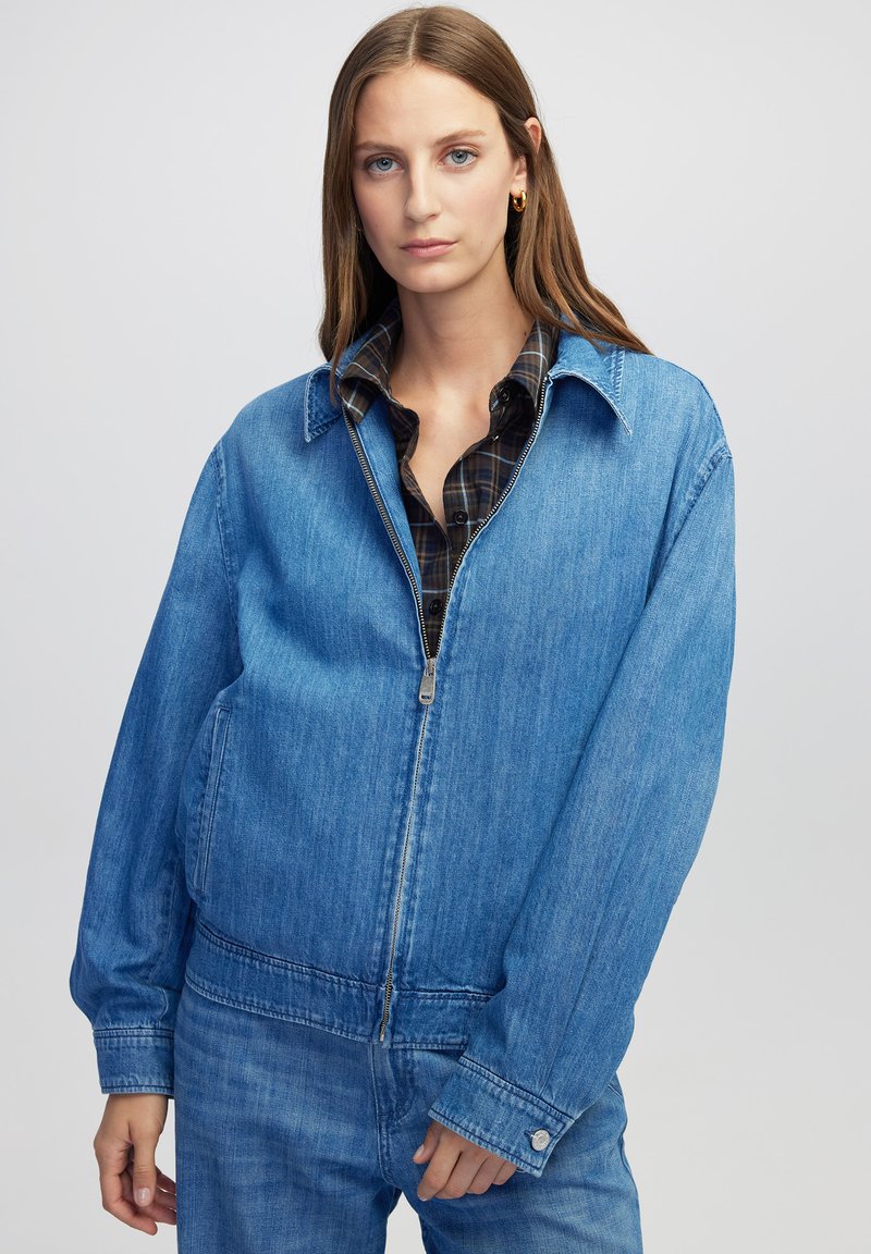 Giovane donna che indossa un ampio giubbotto di denim blu e jeans abbinati, con il davanti parzialmente zipato e una camicia a quadri sotto.