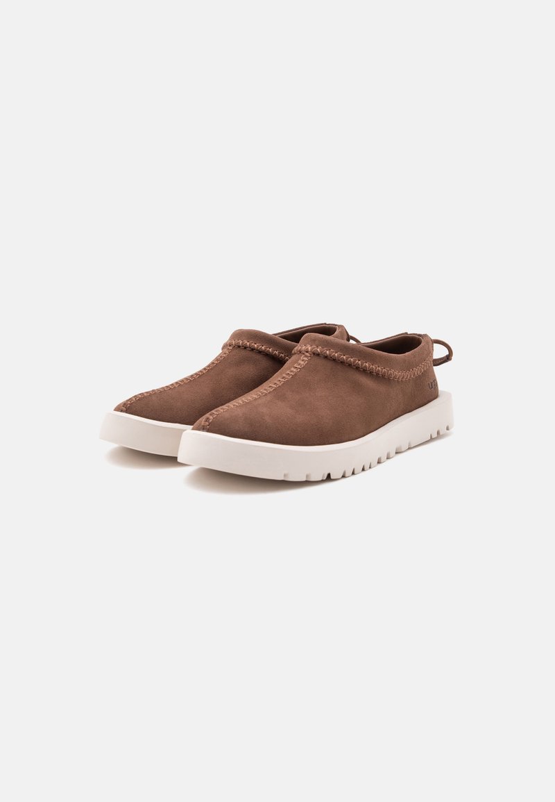 Zapatos casuales marrones sin cordones con suelas de goma blancas y costuras visibles alrededor de los bordes superiores sobre un fondo liso y claro.