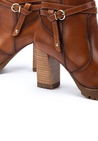 Botas de piel marrón hasta el tobillo con un tacón cuadrado, que presentan una correa y un detalle de hebilla. El tacón tiene una textura de madera estriada.