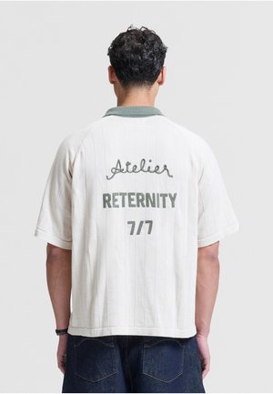 Man met kort krullend haar die een wit shirt met korte mouwen en een groene kraag draagt, met de tekst "Atelier RETERNITY 7/7" geborduurd op de rug.