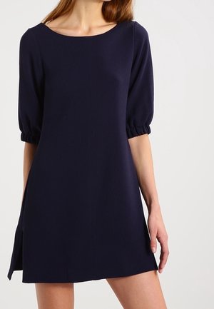 Femme portant une robe bleu marine à manches courtes et longueur genou avec un col bateau et des poignets élastiques, se tenant devant un fond clair.