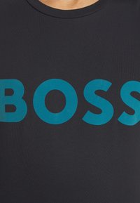 Tmavě modré tričko s kulatým výstřihem, na přední straně velký tyrkysový nápis "BOSS" tištěný tučným písmem bezpatkovým písmem.
