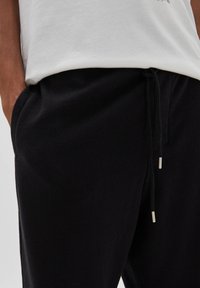 Svarta sweatpants med en mjuk textur, med elastiskt midjeband och dragsko med metallspetsar. Ficka synlig på sidan.