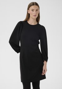 Rochie neagră cu mâneci lungi bufante, decolteu rotund și siluetă dreaptă. Material neted, cu ușoară strălucire și fără modele vizibile.