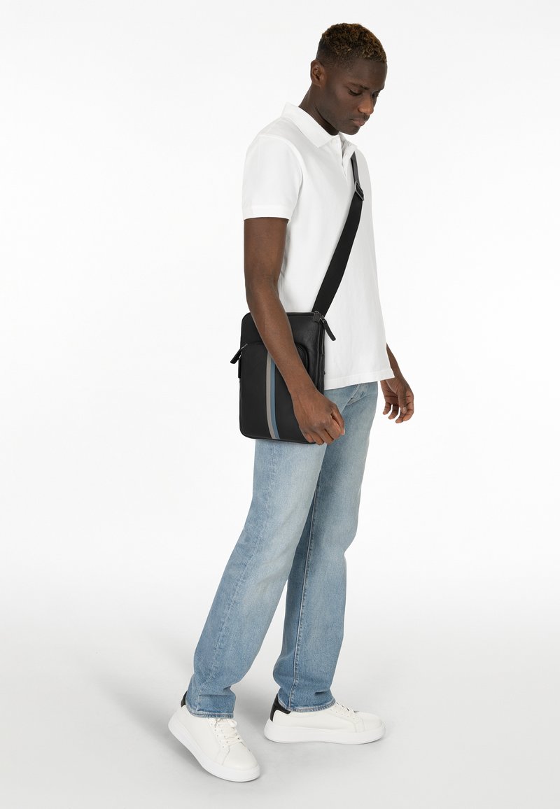 Bolso mensajero sintético negro con dos compartimentos con cremallera, franja de acento azul, usado como bandolera. Una camiseta polo blanca y unos vaqueros azul claro completan el look.