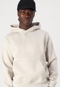 Beige hoodie with a kangaroo pocket, drawstring hood, and ribbed cuffs. Smooth fabric texture, featuring a relaxed fit and minimalist design.