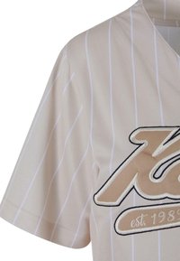 Beige jersej s bielymi pruhmi, vystrih do V a krátkymi rukávmi. Obsahuje vyšívaný text v zlatej a čiernej farbe, čím pridáva textúru a detail.